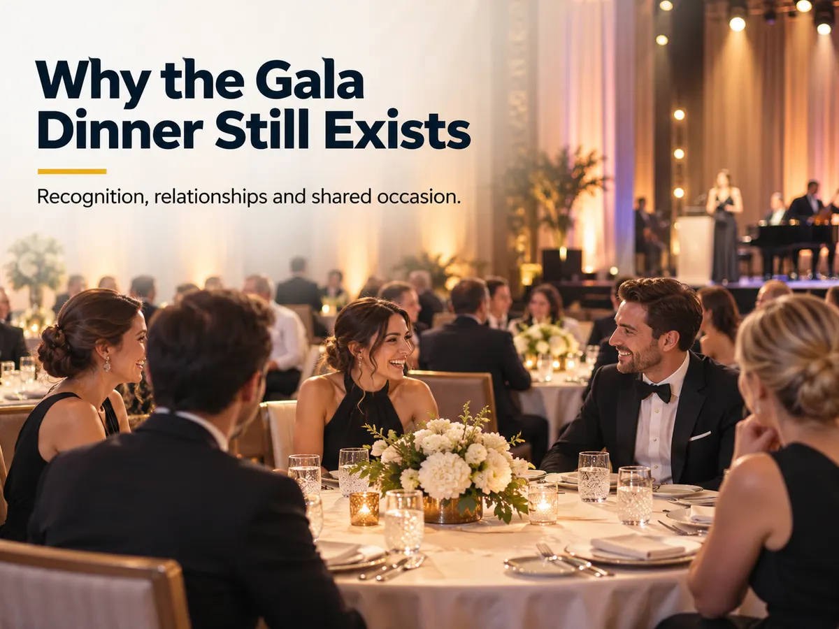 Formally dressed guests talk around a gala dinner table while musicians perform on stage.