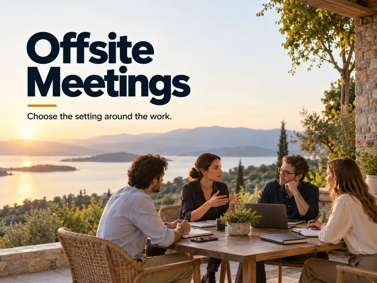 Four colleagues discuss work around a terrace table with notebooks and a laptop overlooking the coast.